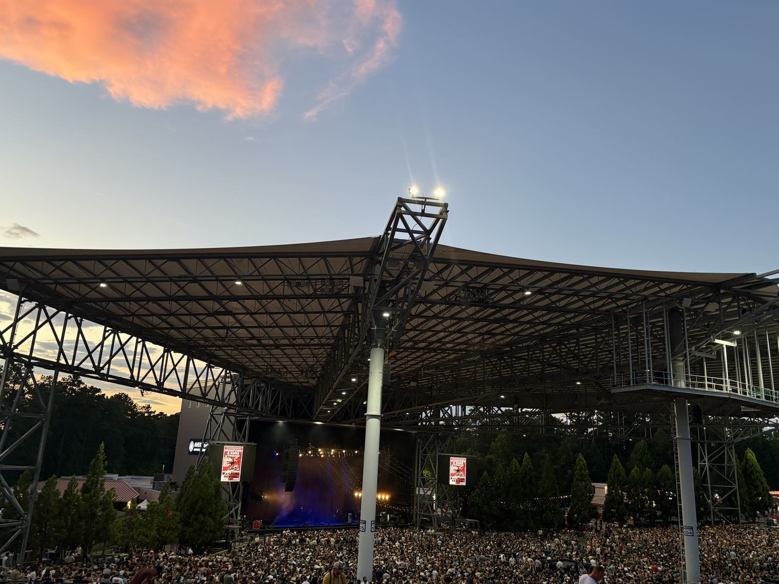 Packed crowd beneath the steel canopy of Ameris Bank Amphitheatre in Alpharetta at sunset, with the stage glowing in the distance