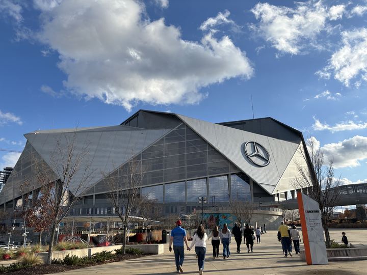 Fans walk toward the angular glass and steel exterior of Mercedes-Benz Stadium on a sunny day